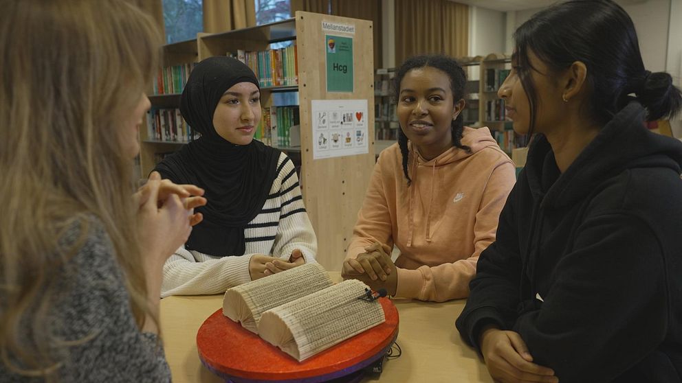 Grupp med fyra personer som sitter runt ett bord i en biblioteksmiljö. På bordet finns en stor bok, och deltagarna diskuterar eller interagerar.