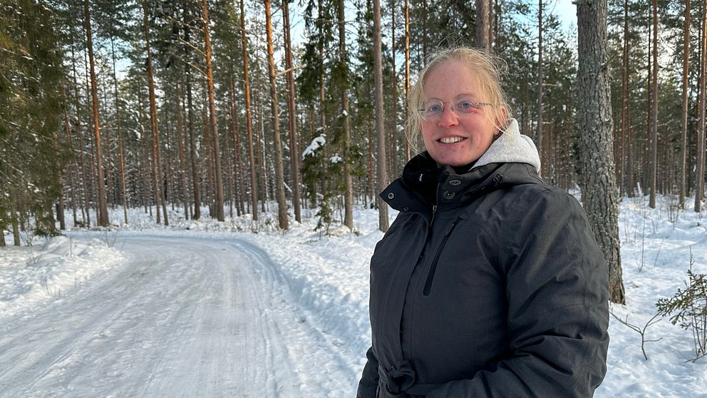 En kvinna klädd i en svart vinterjacka står på en väg i skogen som är täckt av snö. Hon tittar in mot kameran och ler.
