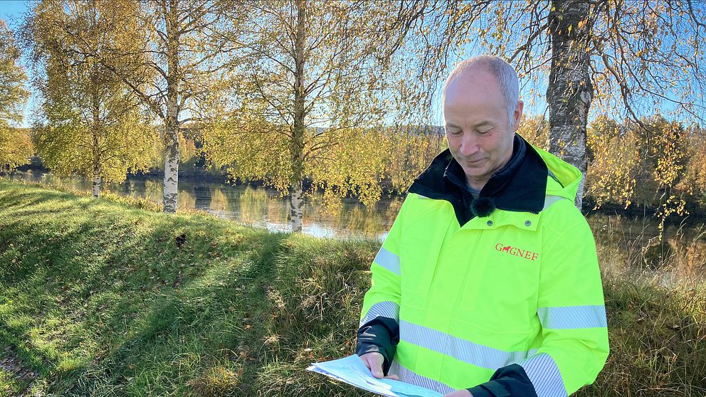 En person i en högsynlighetsjacka står vid en grön gräsyta nära en vattenspegel, med en vall och träd i bakgrunden. Personen håller i en karta.