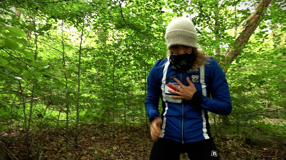 Sara Hagström med mössa, buff och tre jackor i skogen mitt i sommaren.