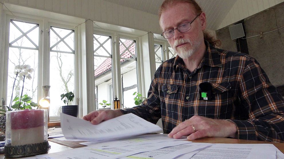Paul Carlbark i rutig skjorta sitter vid ett bord med flera dokument framför sig. Rummet har stora fönster som släpper in naturligt ljus, och det finns inomhusväxter synliga i bakgrunden. På bordet står också ett ljus i en hållare.