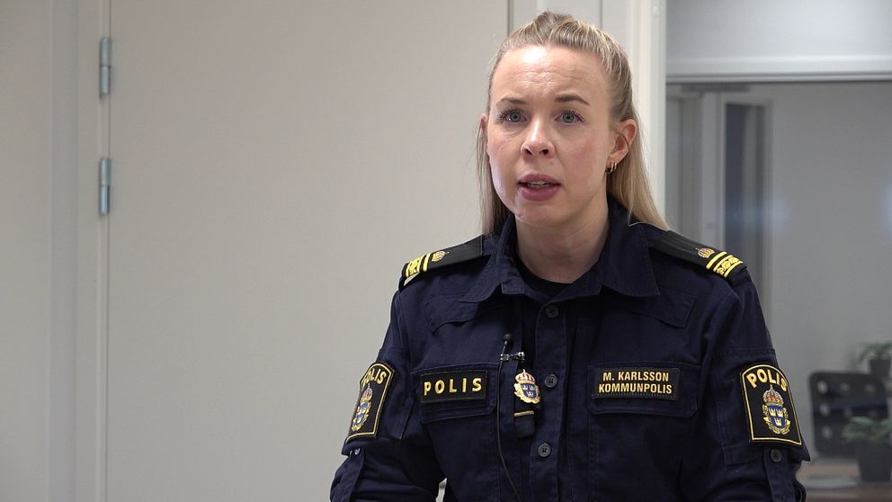 Kommunpolisen Maja Karlsson står och tittar mot kameran i sin mörkblåa polisuniform. Hon har blont hår, i bakgrunden är en vit dörr till vänster om henne och till höger ett fönster.