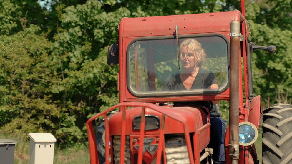 Kvinna kör röd traktor framför grönskande träd i lantlig miljö.