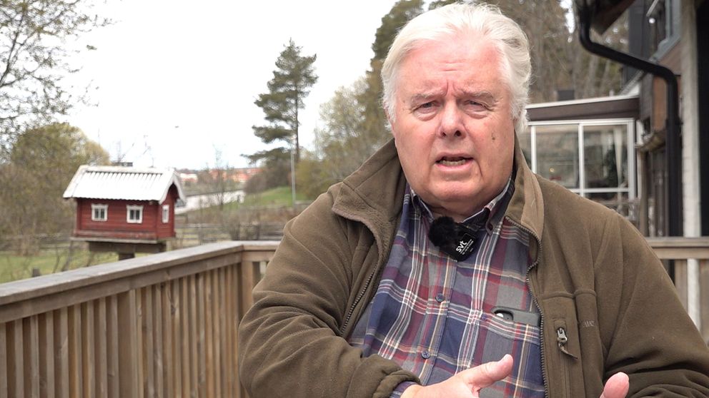 En person i rutskjorta sitter utomhus vid ett trästaket, med en röd fågelholk i bakgrunden.
