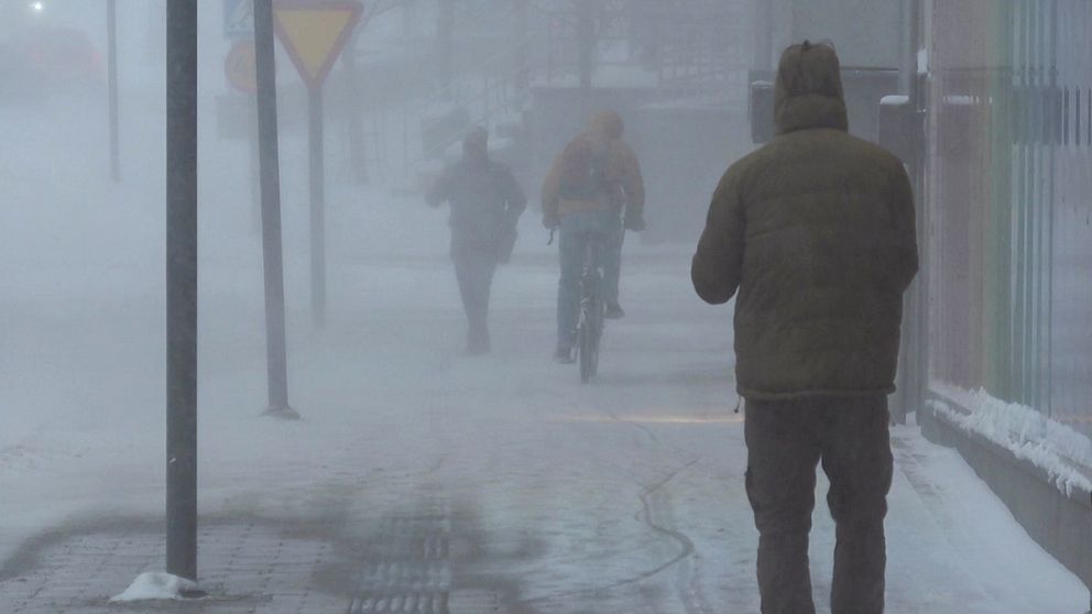 Mörk och snöig stadsmiljö med svag sikt på grund av kraftig snöblandat väder. I förgrunden går en person klädd i en mörk jacka, medan två andra personer, varav en på cykel, syns längre bort i en snöig landskap. Gatuskyltar och byggnader är delvis synliga genom snöverkan.