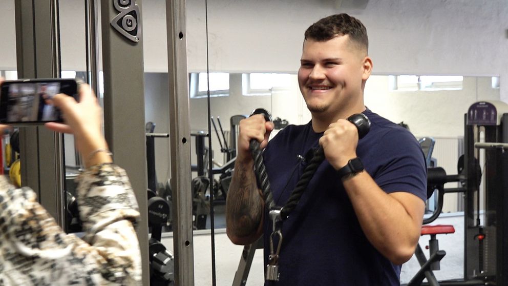En ung man står på ett gym och tränar triceps. Han håller i ett träningsredskap och tittar in i en mobilkamera och ler.