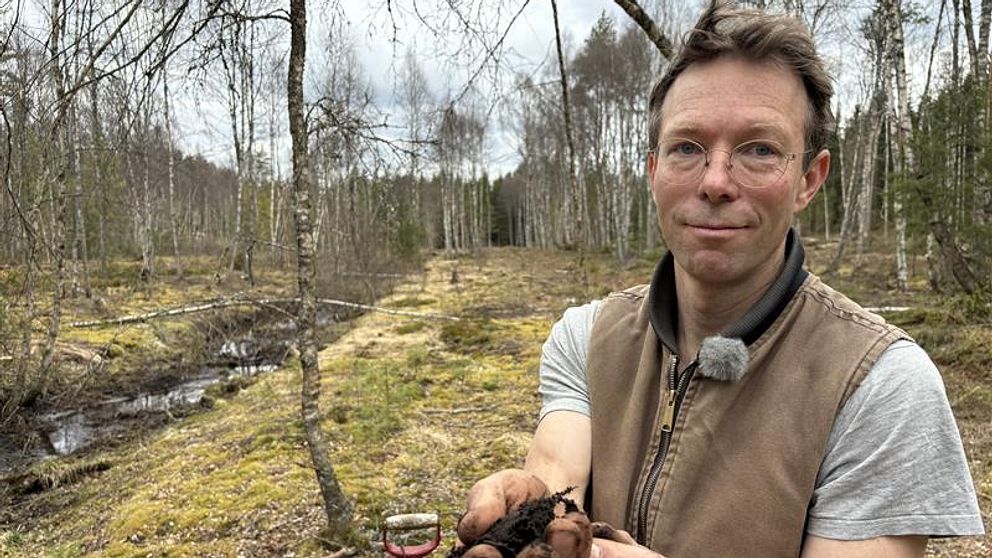 Skogsägaren Simon Jonegård håller i en bunt torv i en skogsmiljö, våtmark, med bäck och mossa.