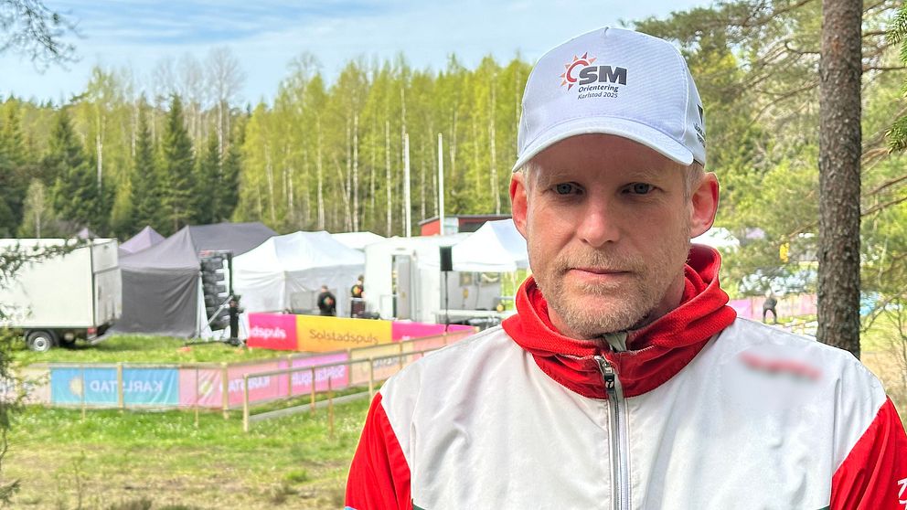 Person i vit GMOK-mössa och jacka vid ett utomhusevenemang i skogsmiljö.
