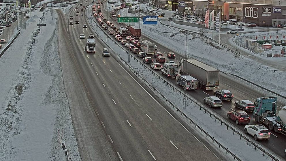 Kö av bilar och lastbilar på en snöbelagd motorväg med flera filer. Den övre delen av bilden visar ett trafikledningssystem och en kommersiell byggnad i bakgrunden.
