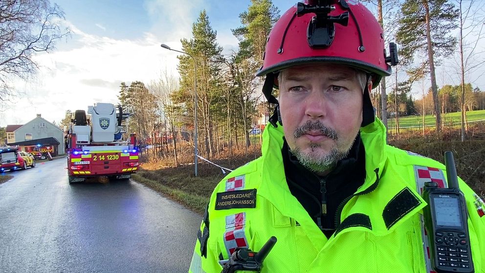 En brandman i en gul och svart skyddsutrustning står framför en räddningsinsats på en väg. I bakgrunden syns en brandbil med blåljus, samt flera andra fordon på platsen. Träden i området ger en naturnära känsla, och det är en klar himmel. Situationen tyder på en aktiv räddningsoperation.
