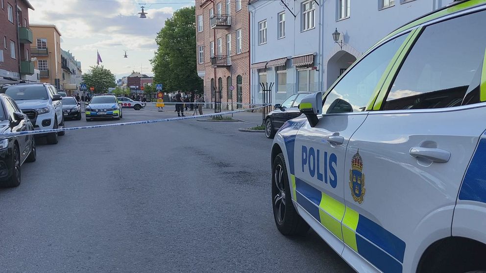 I förgrunden av bilden syns en polisbil. I bakgrunden ser man polisavspärrningar och polis som pratar med folk.