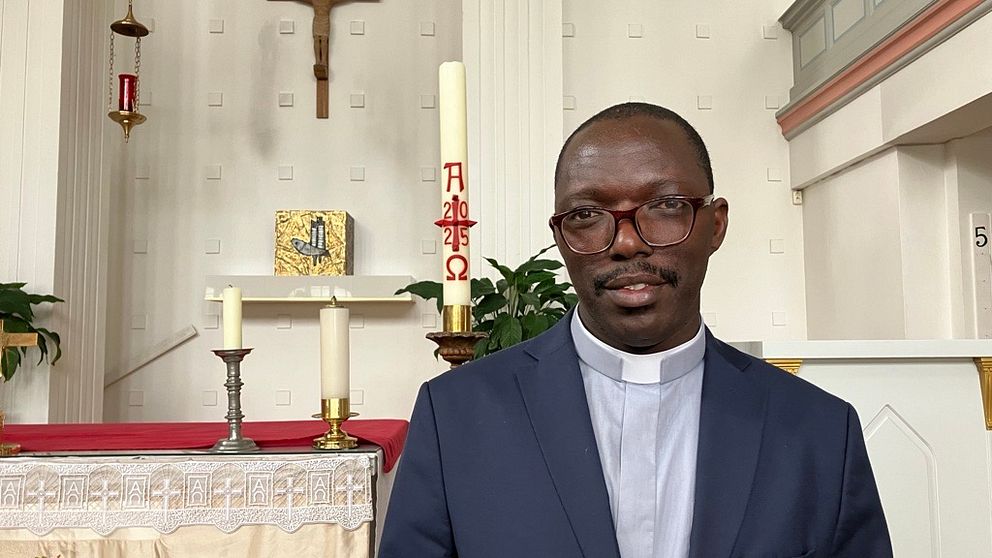 Präst i kavaj står framför altaret i en kyrka med kors och ljus i bakgrunden.