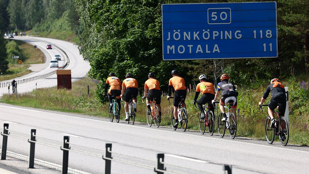 En grupp cyklister i orangea tröjor cyklar längs en väg. Bakom dem syns en vägskylt som visar avståndet till Jönköping (118 km) och Motala (11 km). I bakgrunden är en grönskande skog och en bilväg synlig.