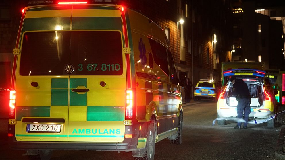En ambulans står parkerad på en mörk gata, med ljusen tända. En person syns i närheten av ambulansens baklucka, vilket tyder på en pågående situation. Bakom ambulansen syns en annan bil, kanske i färd med att assistera. Gatan är svagt upplyst av omgivande byggnader.