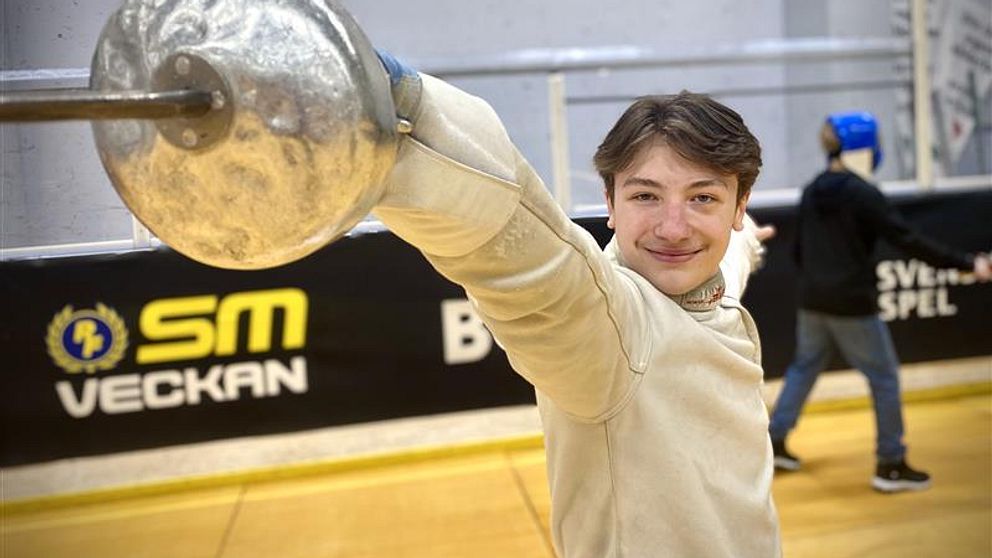 Vincent Ryde, SM-veckan i Borås 2025. En fäktare i vit dräkt sträcker fram ett svärd mot kameran med fokus på bladet. I bakgrunden finns en annan fäktare i skyddsutrustning. En banderoll med texten ”SM Veckan” kan ses på väggen bakom.