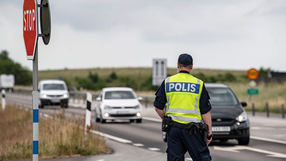 En polis står vid en väg med en STOPP-skylt som syns i bakgrunden. Flera bilar närmar sig, och polisen övervakar trafiken. Himlen är molnig och landskapet runt omkring är grönt och öppet.