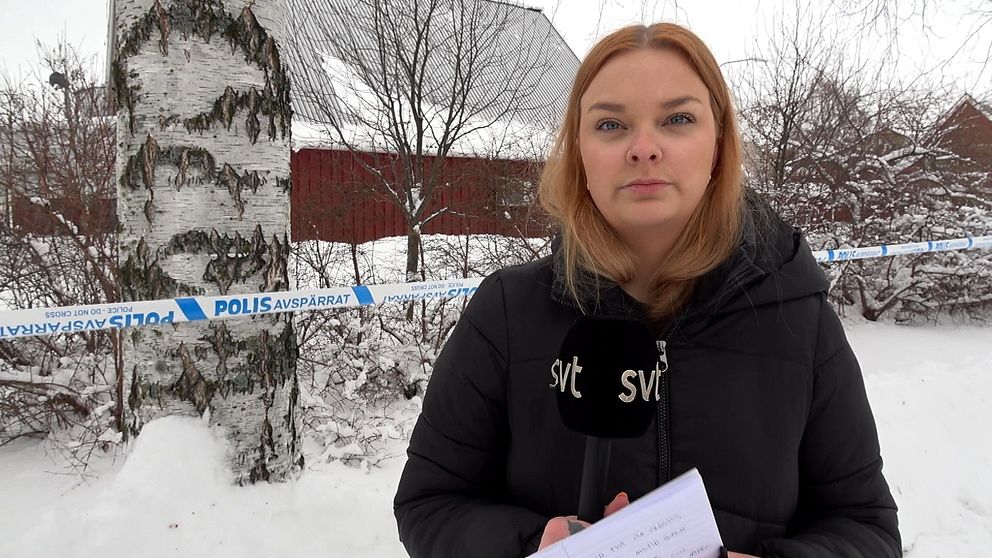 En reporter från SVT står utomhus med en mikrofon i handen, bredvid ett område som har avspärrats av polis. I bakgrunden syns en snötäckt björk och några byggnader.