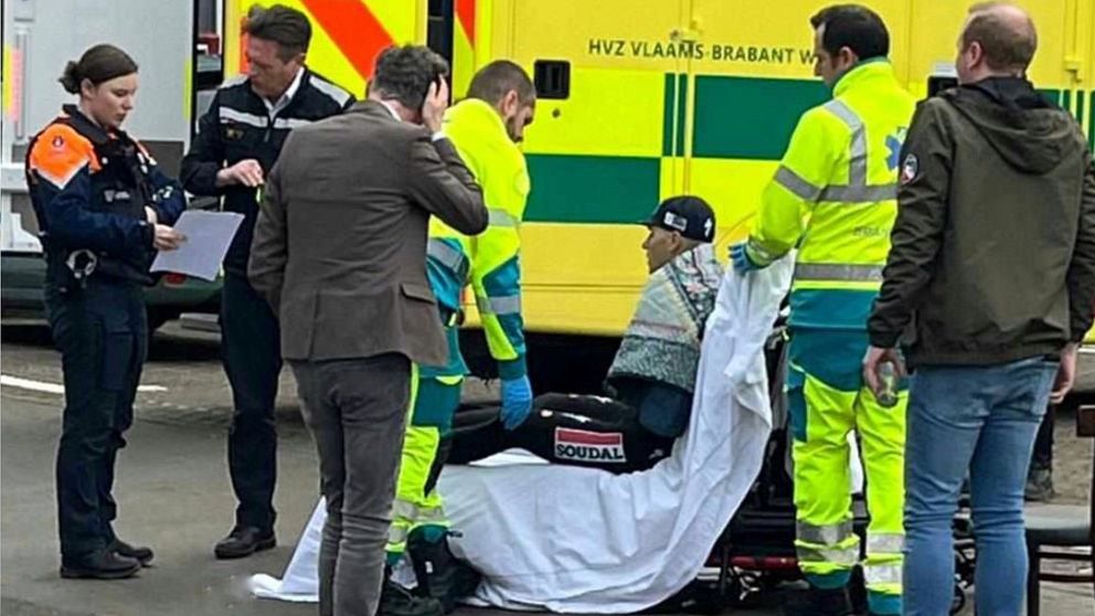 En grupp räddningspersonal assisterar en skadad person på en bår, täckt med en vit filt. Två av räddningsarbetarna har på sig reflexkläder och en tredje bär en jacka med logotyp. I bakgrunden syns en ambulans med tydliga märkningar. Enligt situationen förefaller det vara ett akut ärende, med flera personer närvarande, inklusive en polis.