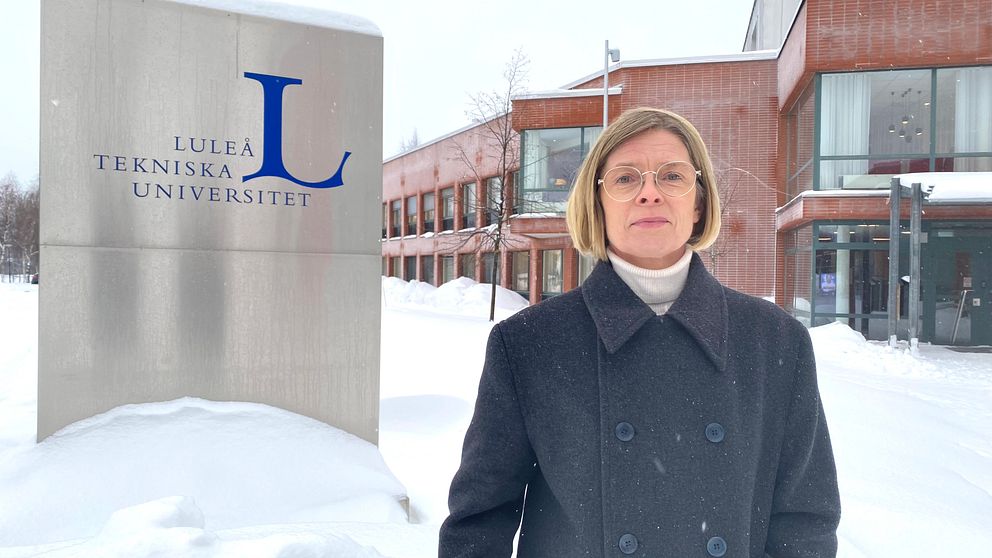 En person står framför en skylt för Luleå tekniska universitet, omgiven av snö, med universitetsbyggnader i bakgrunden.