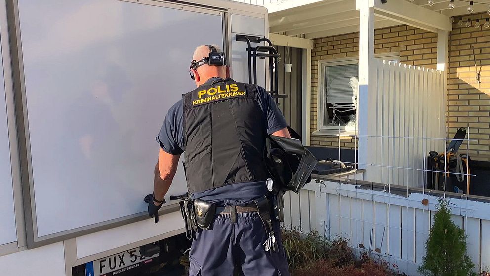En polis på en brottsplats arbetar vid en bil. I bakgrunden syns en bostad med ett trasigt fönster.
