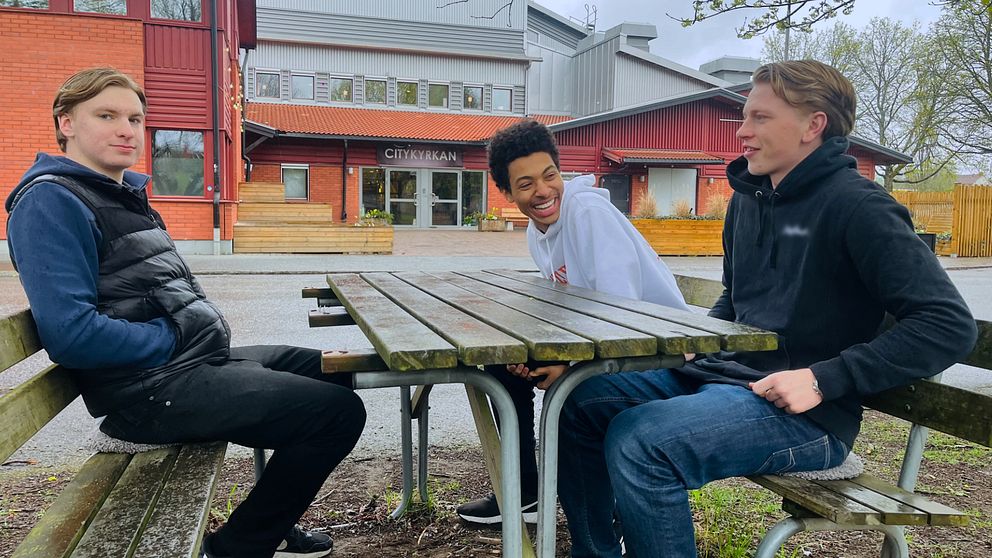 Tre personer sitter vid ett utomhusbord framför en byggnad. En av dem skrattar.
