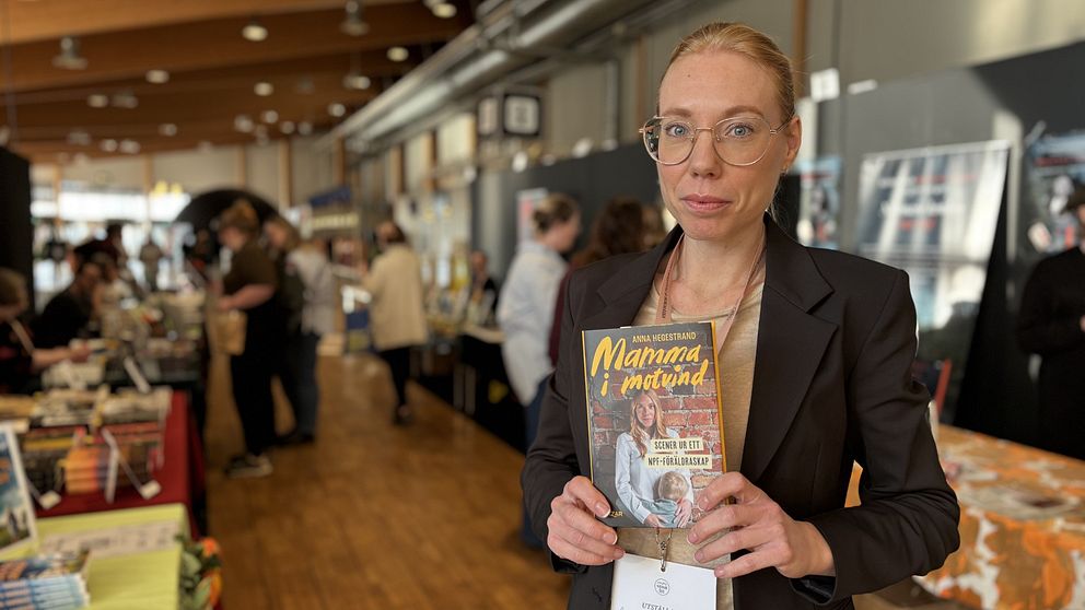 En person håller upp en bok med titeln ”Mamma i mittend” på en bokmässa där människor bläddrar och samtalar vid olika bokstånd i bakgrunden.