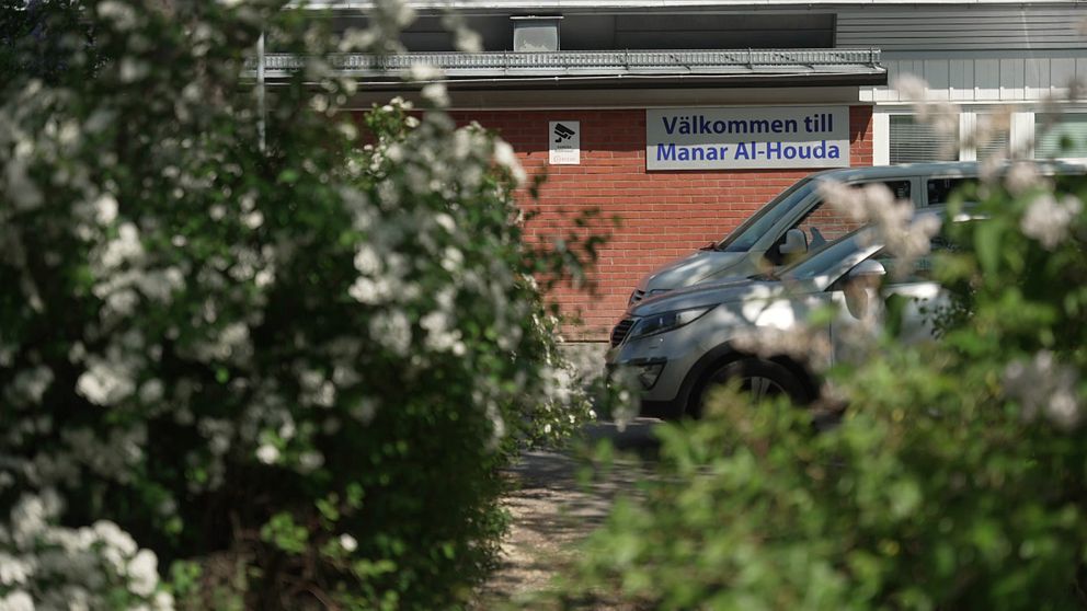 Buskage i förgrunden. Längre bort står bilar parkerade och skolans skylt,