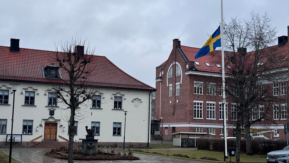 Två äldre byggnader i en tätort under en gråmulen himmel, med svenska flaggan hissad på halv stång, vilket ofta indikerar sorg eller ett officiellt sorgebesked.