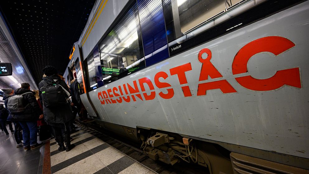 En bild av en tågplattform där resenärer väntar på att stiga på ett tåg markerat med ”ÖRESUNDSTÅG”. Tåget står stilla vid plattformen och det finns en tydlig utskrift av logotypen på sidan av tåget. Resenärerna är klädda för vintern, och stationens interiör är synlig i bakgrunden.