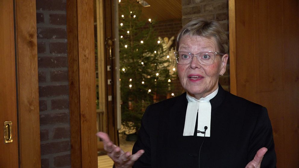 En person i prästkläder gestikulerar medan de pratar. I bakgrunden syns en gran prydd med ljus, vilket ger en festlig atmosfär. Rummet har en varm och inbjudande känsla med synliga träpaneler och mjukt ljus.