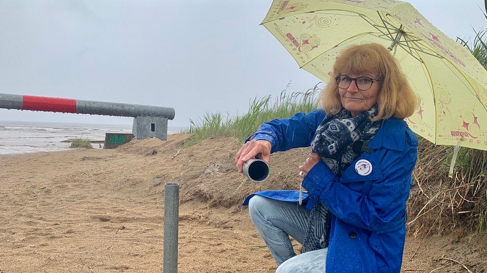 Eva Hallberg sitter på knä under ett paraply framför en vägbom på en strand som sågats av.