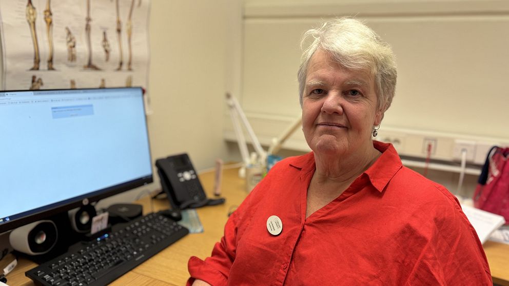 En äldre person med vitt hår sitter vid ett skrivbord i ett kontor, iklädd en röd skjorta. På skrivbordet står en datorskärm, tangentbord och telefon. På väggen bakom finns en plansch med anatomiska inslag.
