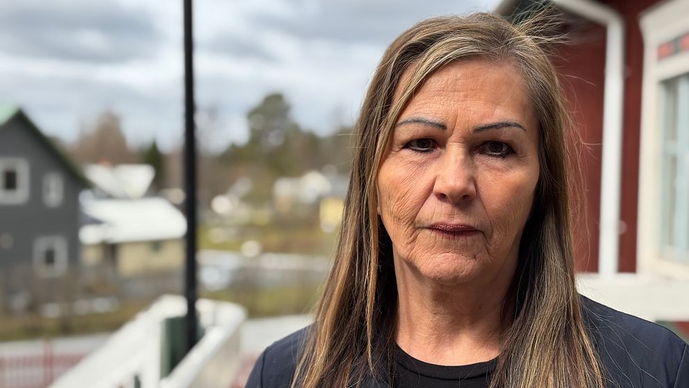 Kvinna med långt hår står utomhus vid ett hus, bostadsområde syns i bakgrunden.