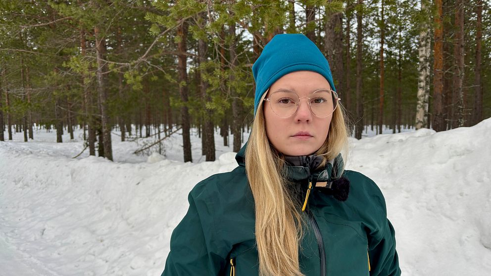 En person klädd i mörk jacka och blå mössa står i ett snöigt skogslandskap med tallar i bakgrunden.