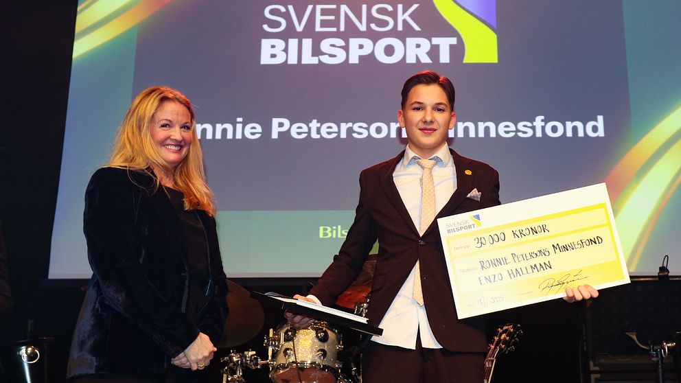 Enzo Hallman i kostym tar emot en stor check på 30 000 kronor av Nina Kennedy under en prisutdelning för Svensk Bilsport. I bakgrunden syns trummor och en skärm med texten: ”Ronnie Petersons Minnefond”.