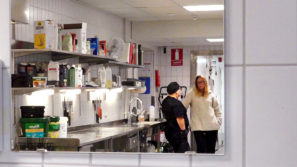 Två personer samtalar i en lukas köksmiljö, med en rad köksredskap och förvaringsbehållare bakom dem. Den ena personen står vid en disk, medan den andra står i närheten och lyssnar. Rummet är upplyst och ser välorganiserat ut.