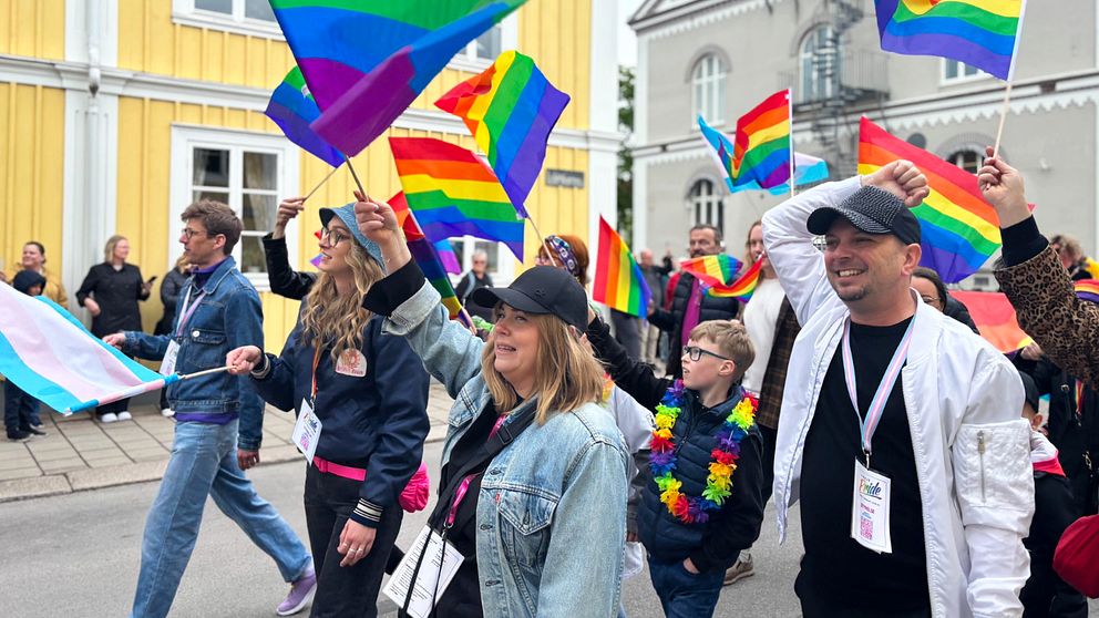 Deltagare går i prideparad iVäxjö och vinkar med regnbågsflaggor på en stadsgata.