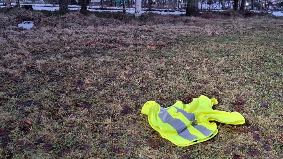 En gul västar med reflexer ligger på marken i ett grönskande område med viss brun jord och några grässtrån, omgivna av träd.