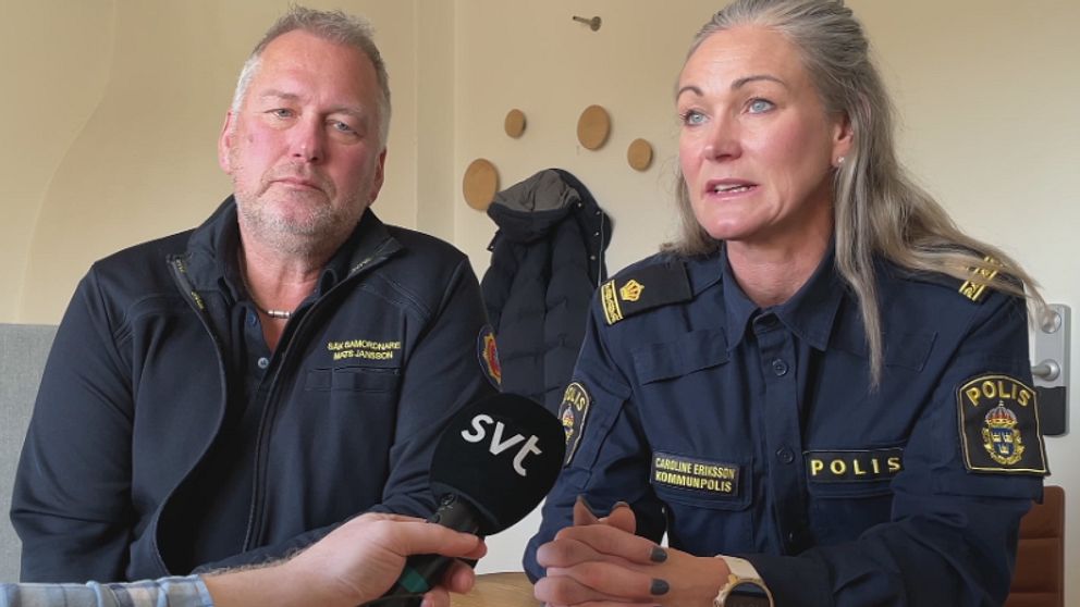 En intervju sker mellan två personer, där en person bär en polisuniform med märket ”POLIS” och den andra har en jacka med texten ”SÄKERHETSANSVARIG.” En mikrofon hålls i bild, som visar att det handlar om en medieintervju. Bakgrunden har en enkel inredning och en jacka hänger på väggen.