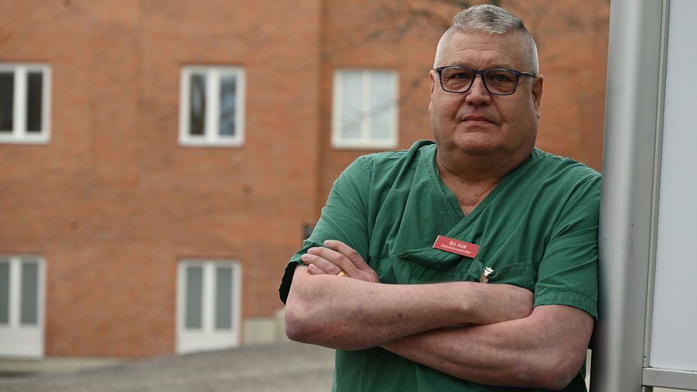Man i grön arbetskläder står med korslagda armar framför en tegelbyggnad.
