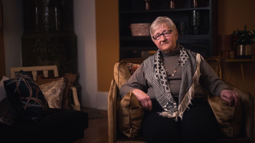 En person sitter i en mjukt belyst, hemmasittande miljö. Hon har på sig en scarf och en tröja, och är omgiven av dekorativa kuddar och en bokhylla i bakgrunden. Rumskänslan är varm och inbjudande.