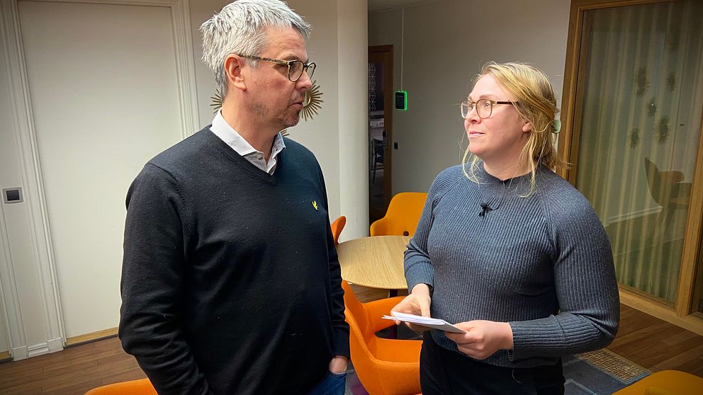 Två personer står mittemot varandra i ett modernt inomhusmiljö med orange stolar och ett runt bord. Den ena personen håller i papper och har på sig en grå tröja, medan den andra bär en svart tröja.