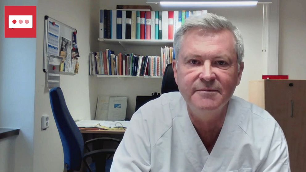 En läkare i vit laboratoriejacka sitter framför en dator. I bakgrunden syns en bokhylla fylld med böcker och några dokument på ett skrivbord. Rummet är ljust och professionellt inrett, vilket skapar en arbetsmiljö.