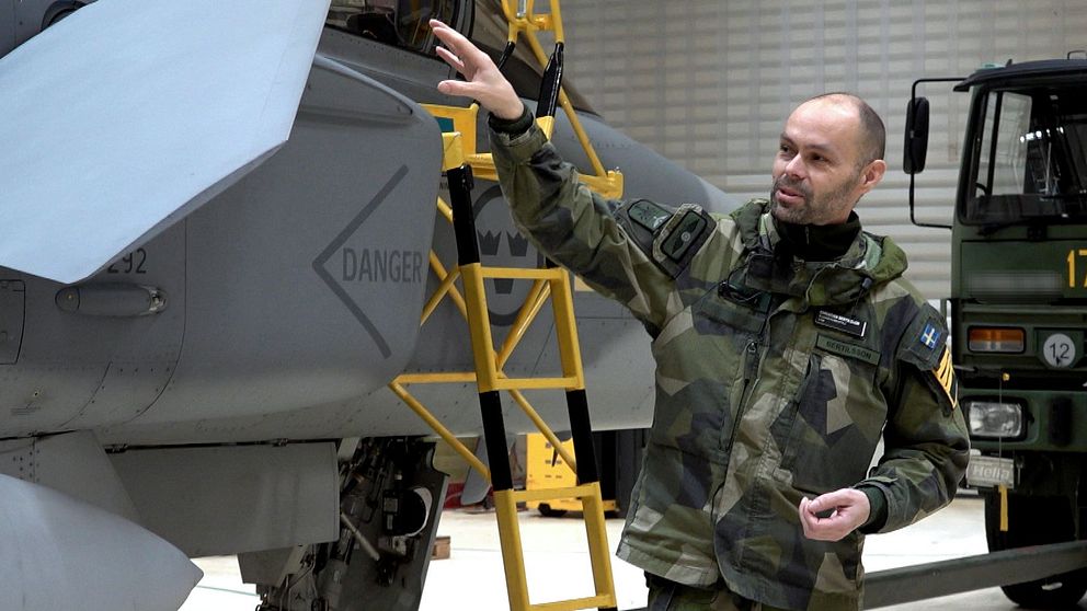 En person i militäruniform pekar mot en del av ett flygplan inuti en hangar, med en stege placerad intill. I bakgrunden syns en militär lastbil och andra detaljer från hangaren.