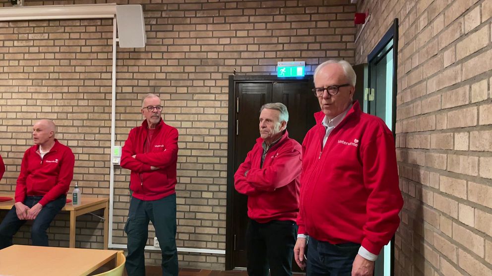 Fyra personer i röda jackor står i en inomhusmiljö med tegelväggar. Belysning kommer från taklampor och en dörr syns till vänster i bilden.