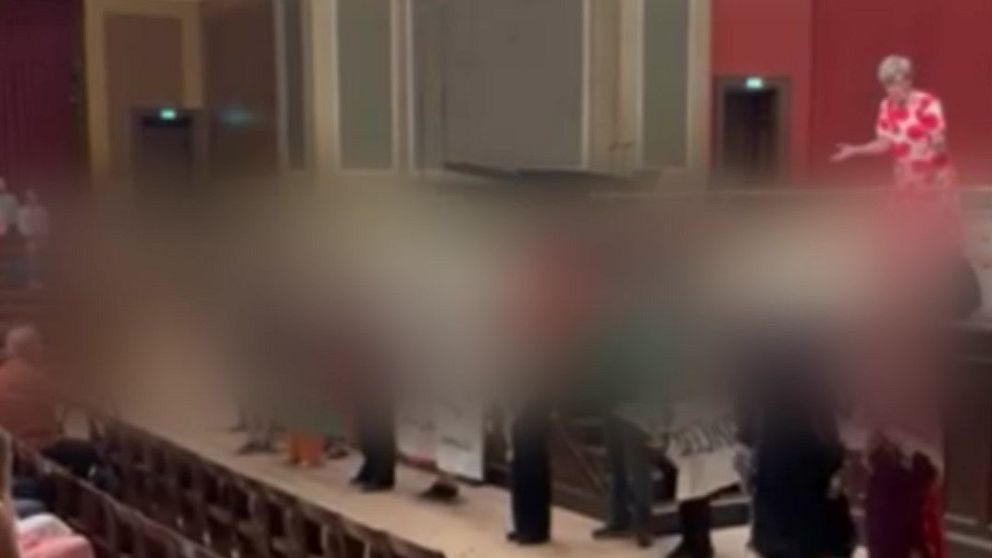 En scen i en teater med en person i röd blommig klänning som talar, medan flera personer står framför med en banner.