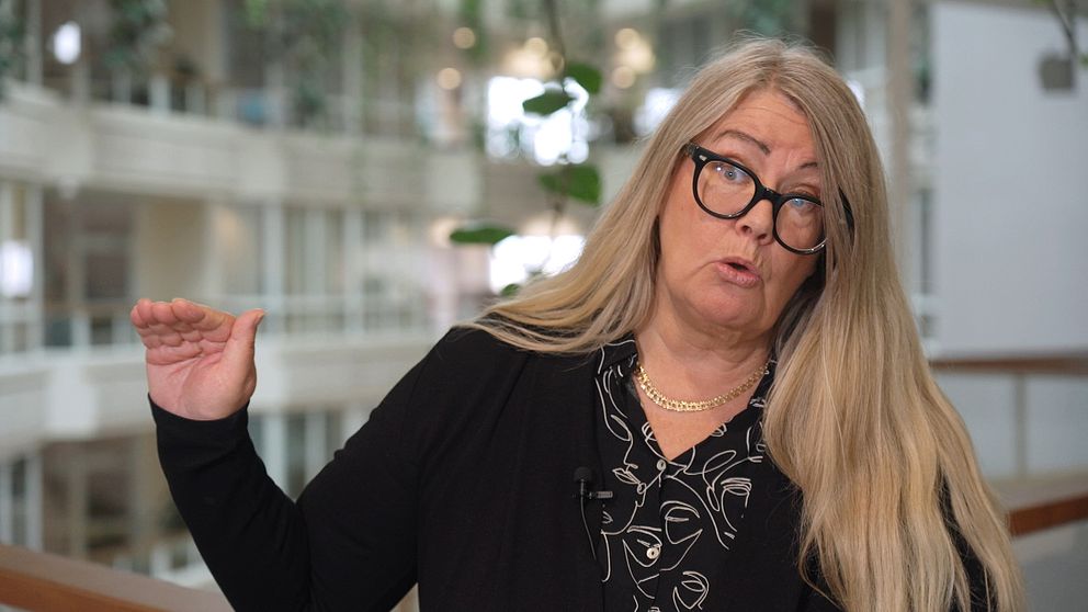 Annika Wallenskog med långt, blont hår gestikulerar med sin hand i en inomhusmiljö. Bakgrunden visar en modern byggnad med stora fönster och växtlighet, vilket skapar en ljus och öppen atmosfär. Personen är klädd i en svart tröja med mönster.
