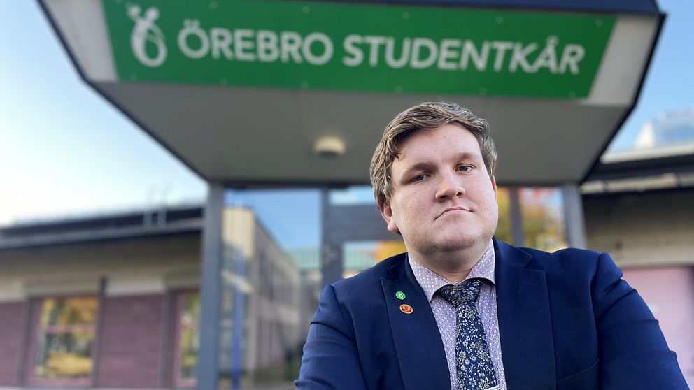 En person i kostym står framför Örebro Studentkårs lokaler. Byggnaden har en tydlig grön skylt med namnet.