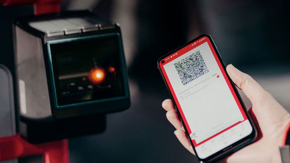 En person håller upp en smartphone med en QR-kod mot en QR-kodsläsare. Skärmen på telefonen visar en app med QR-koden och tillhörande information. Bakgrunden är suddig och fokuserar på interaktionen mellan telefonen och läsaren.