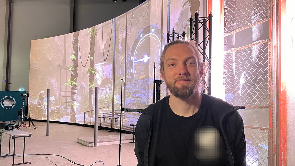 En man står framför en stor,  rundad bildskärm med projektioner av en grönskande, drömlik scen. Rummet är inrett med olika strukturer som stödjer ljus- och ljudutrustning, och det finns ledningar på golvet som tyder på en pågående produktion eller inspelning.